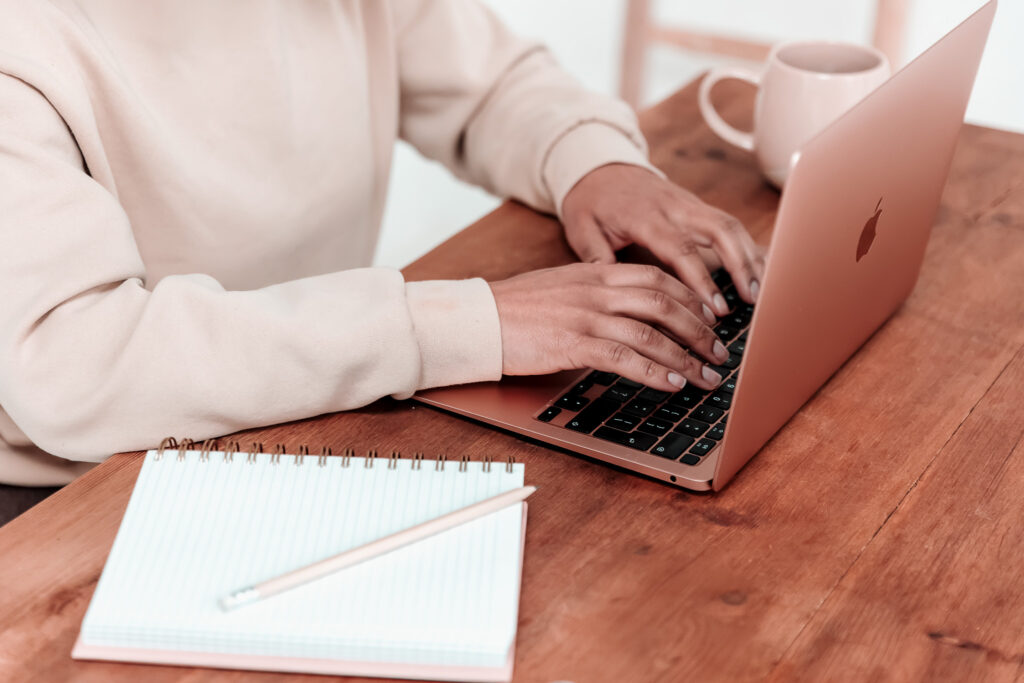 A woman sat at an apple lapop, fingers are positioned as if they are typing. On the table beside her is a blank spiral bound notebook with a pencil on the top.